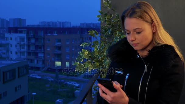 Молодая, красивая женщина стоит вечером на балконе и работает на смартфоне — стоковое видео