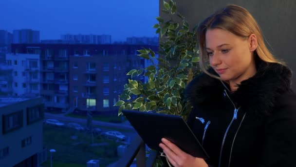 Μια νεαρή, όμορφη γυναίκα στέκεται σε ένα μπαλκόνι το βράδυ και λειτουργεί σε ένα δισκίο — Αρχείο Βίντεο