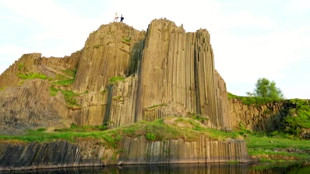 People stand on the top of a columnar basalt rock near a lake, the bright sky in the background — Stock Video