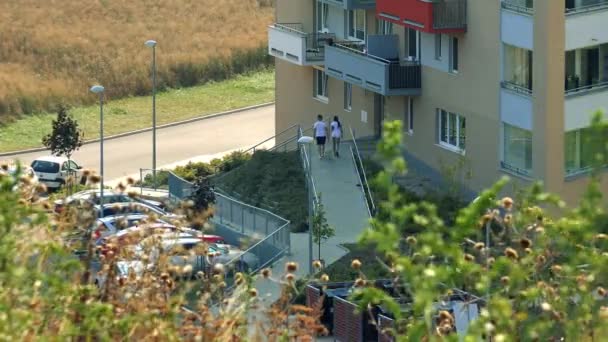 Menschen betreten ein Mehrfamilienhaus, niedrige Vegetation im Hintergrund, ein kleiner Parkplatz neben dem Gebäude, Gras und ein Baum im Vordergrund - Draufsicht — Stockvideo