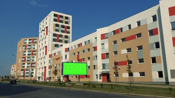 En skylt med en grön skärm av en väg i ett stadsområde, bilar förbi, färgglada flerbostadshus och klarblå himmel i bakgrunden — Stockvideo