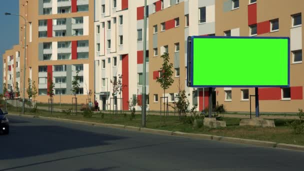 Билборд с зеленым экраном на дороге в городской местности, проезжают машины, красочные жилые дома на заднем плане — стоковое видео