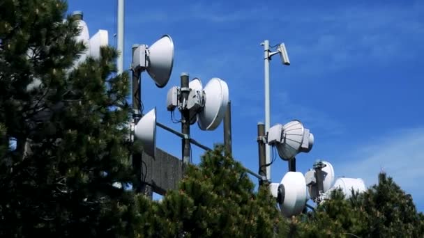 Parabol antenner av en television tower på ett räcke underifrån, den ljusa blå himmel i bakgrunden, trädkronorna i förgrunden — Stockvideo