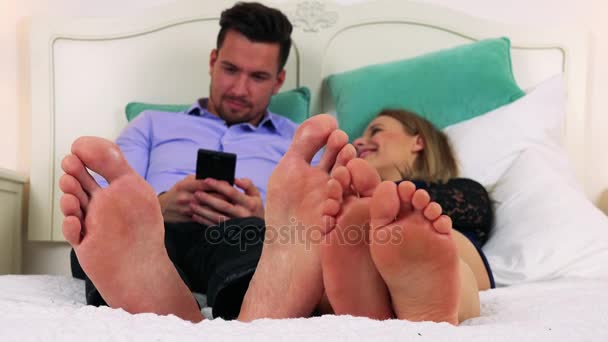 Un hombre y una mujer yacen en una cama con un smartphone — Vídeo de stock
