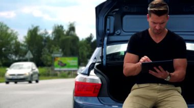 adam araba bagajında oturur ve tablet üzerinde çalışır