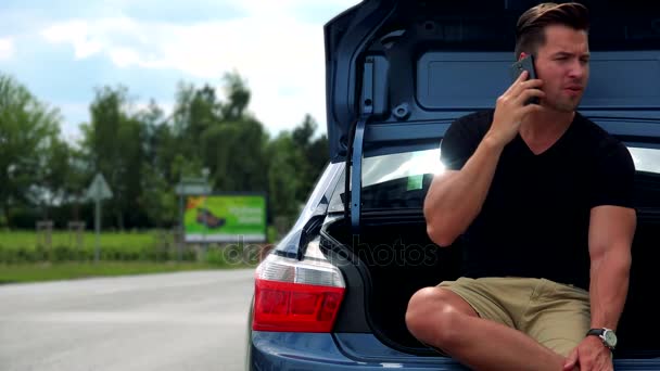 男は車のトランクに座っているし、スマート フォンと携帯電話 — ストック動画