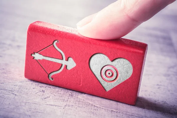 Cupidos arco y flecha apuntando a un corazón grabado en un bloque de madera roja y arreglado por un dedo femenino en una mesa — Foto de Stock