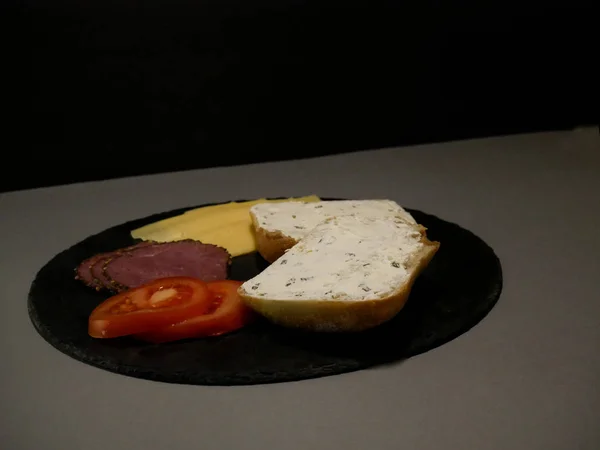 Pão fatiado, queijo e presunto em prato preto e fundo cinza — Fotografia de Stock