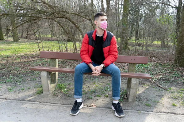 Портрет брюнетки мужчины в хирургической повязке, сидящего в парке на скамейке запасных, коронавирус, болезнь, инфекция, карантин, медицинская маска — стоковое фото