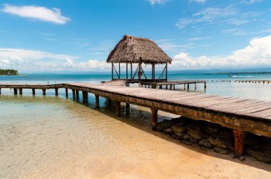Mavi denizde, su üzerinde ahşap yolu ve mavi gökyüzü olan bir kulübe.