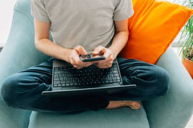 Akıllı telefonda dizüstü bilgisayarı olan bir adam evde turkuaz koltukta turuncu yastıkla çıplak ayakla çalışıyor.