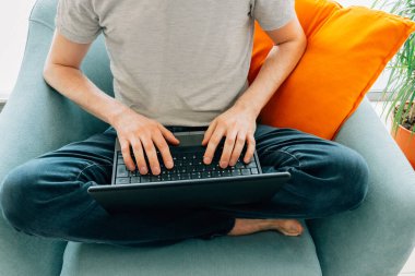 Evde dizüstü bilgisayarla çalışan bir adam turkuaz koltukta çıplak ayakla turuncu yastıkla oturuyor.