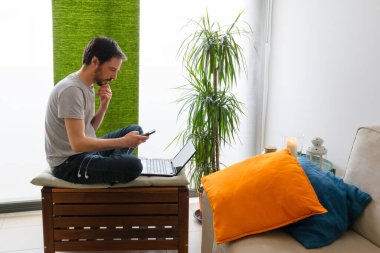 Akıllı telefona bakan adam, tahta taburede yalın ayak, renkli minderlerle oturuyor.