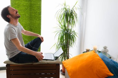 Video konferansında gülen bir adam çıplak ayakla renkli minderlerle tahta taburede oturuyor.