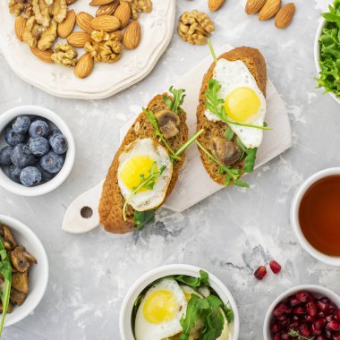 Sağlıklı kahvaltı ile kızarmış yumurta, taze ekmek, salata, çilek, domates, fındık, içecek Kupası, roka, gri hafif bir arka plan üzerinde mantar. Üstten Görünüm