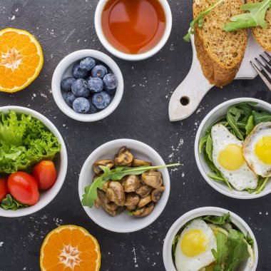 Karanlık bir arka plan üzerinde Kahvaltı: kızarmış bıldırcın yumurtası, roka, taze ekmek, marul, domates, portakal, fındık, çay, kahve, mantar, yaban mersini. Üstten Görünüm