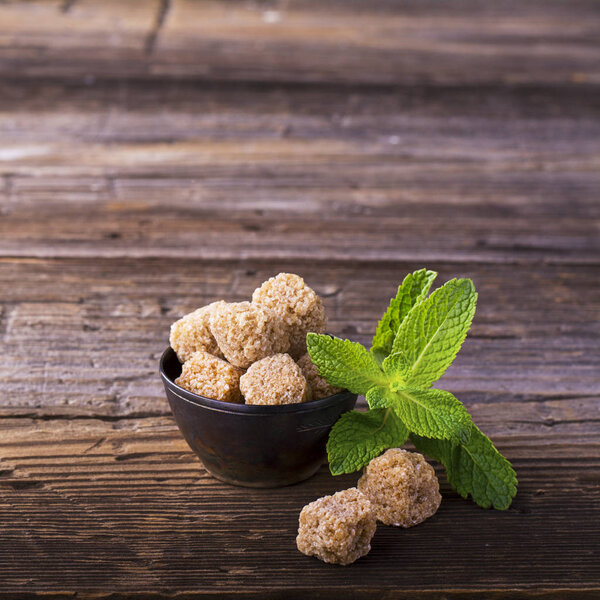 Lump brown cane sugar in a vintage metal dishes with sprig of fresh mint on simple wooden background. The concept natural organic food.
