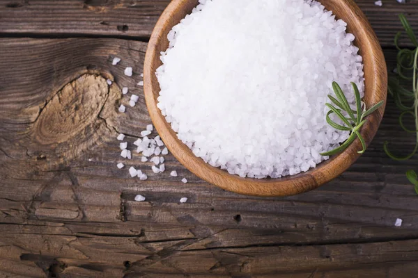 Large White sea salt in a natural wooden bowl with  sprig of fresh rosemary on  simple  background. Selective focus. — Stock Photo, Image