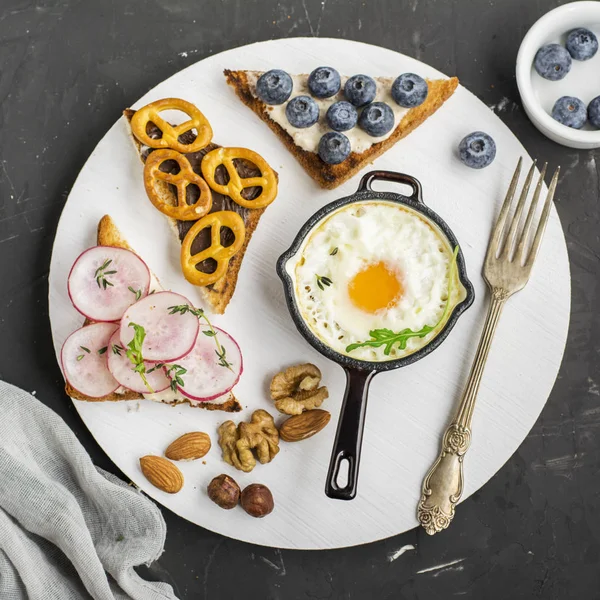 Tasty modern breakfast. Fried eggs in a batch cast iron skillet and toast platter with peanut butter, blueberries, radishes  pretzels cookies on the board   dark background. Top view