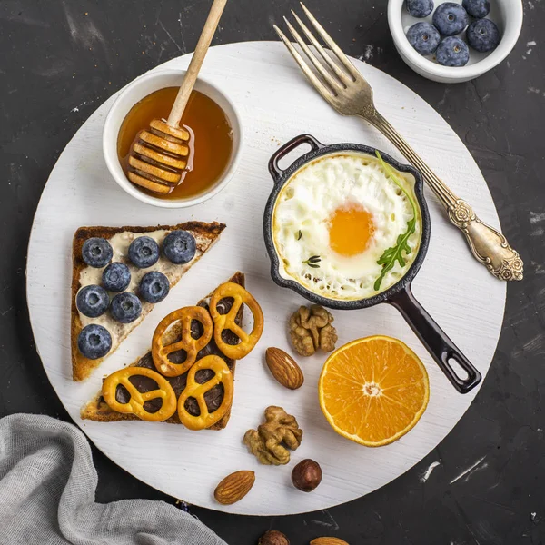 Tasty modern breakfast. Fried eggs in a batch cast iron skillet and toast platter with peanut butter, blueberries, radishes  pretzels cookies on the board   dark background. Top view
