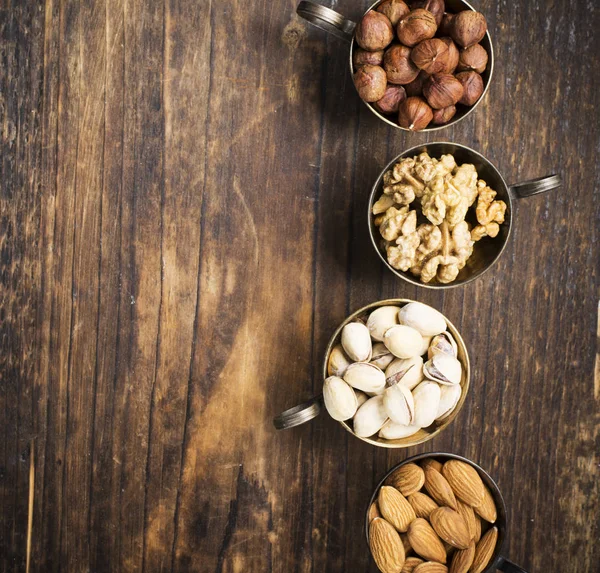 Cuatro copas de cuproníquel vintage llenas de diferentes frutos secos, almendras, pistachos, nueces y avellanas sobre un fondo de madera oscura. Vista superior —  Fotos de Stock