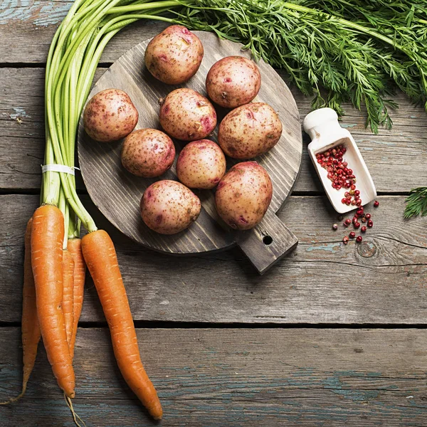 Fresh organic farm vegetables: juicy carrots, young potatoes on a simple wooden background with spices and a knife. Top View. — Stock Photo, Image