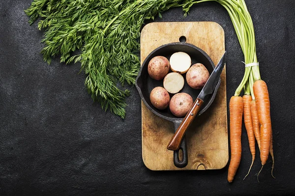 Fresh organic farm vegetables: carrots, potatoes, fresh herbs, spices and oil on a dark textured background with a vintage cutting board and knife. Top View. — Stock Photo, Image