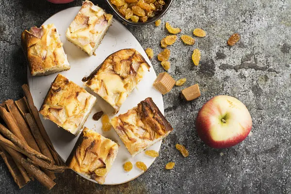 A simple homemade pie with apples and sour cream on a gray background with cinnamon sticks, an apple, caramel sweets. Top view. Selective focus. — Stock Photo, Image