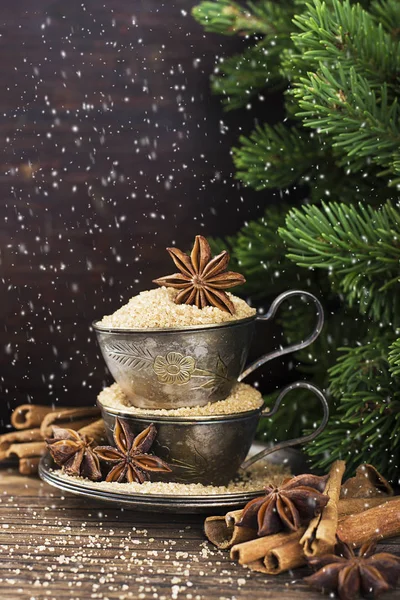 Melchior vintage cups with cane sugar, anise stars, Indian Indian cinnamon sticks on an aged wooden background surrounded by spruce spruce branches. Toned, with imitation of falling snow. Selective fo — Stock Photo, Image