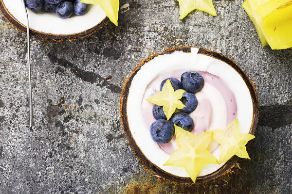 Een gezond ontbijt snack in helften van kokosnoot van berry yoghurt van twee kleuren met bosbessen en carambola's op een eenvoudige grijze stenen achtergrond. Bovenaanzicht. — Stockfoto
