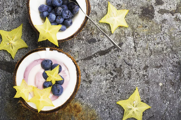 A healthy breakfast snack in halves of coconut from berry yogurt of two colors with blueberries and carambola on a simple gray stone background. Top View. — Stock Photo, Image