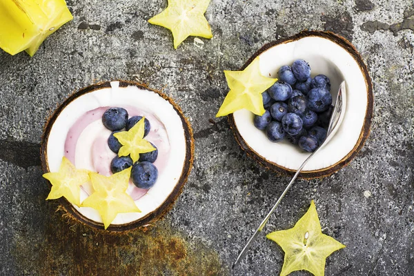 Un saludable snack de desayuno en mitades de coco de yogur de bayas de dos colores con arándanos y carambola sobre un simple fondo de piedra gris. Vista superior . —  Fotos de Stock