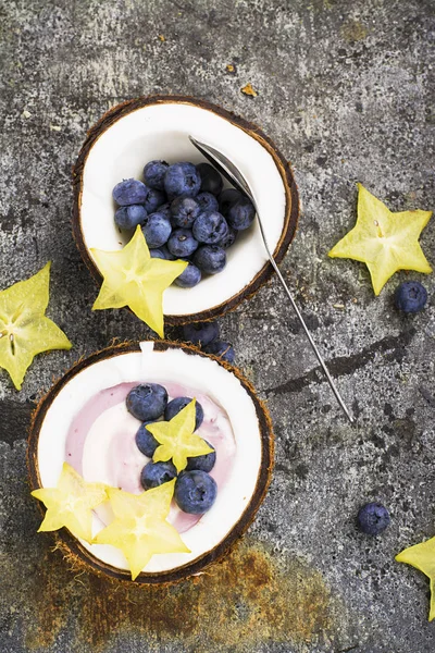 A healthy breakfast snack in halves of coconut from berry yogurt of two colors with blueberries and carambola on a simple gray stone background. Top View. — Stock Photo, Image