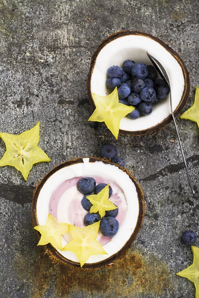 Een gezond ontbijt snack in helften van kokosnoot van berry yoghurt van twee kleuren met bosbessen en carambola's op een eenvoudige grijze stenen achtergrond. Bovenaanzicht. — Stockfoto