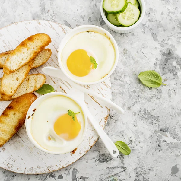 Breakfast for two. Eggs baked in the oven in ceramic portioned forms with toasted bread and slices of fresh cucumber. Top View. — Stock Photo, Image