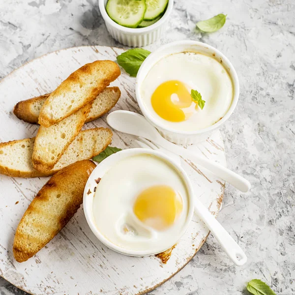 Breakfast for two. Eggs baked in the oven in ceramic portioned forms with toasted bread and slices of fresh cucumber. Top View. — Stock Photo, Image