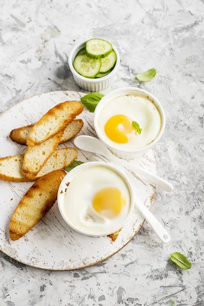 Breakfast for two. Eggs baked in the oven in ceramic portioned forms with toasted bread and slices of fresh cucumber. Top View. — Stock Photo, Image