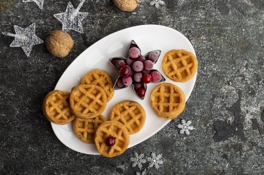 Gümüş Noel dekor ve çelenk bir gri grunge zemin üzerine beyaz oval seramik tabak üzerinde organik Kuzey kızılcık, dondurulmuş çilek ile kahvaltıda ev yapımı yumuşak waffle. Üstten Görünüm.
