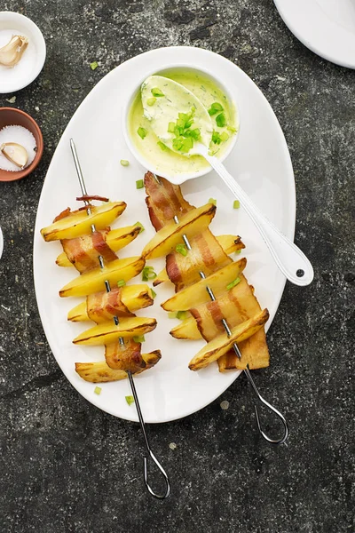 Аппетитные шашлыки с картофельными ломтиками в ломтиках бекона, запеченные в духовке, подаются с йогуртом и зеленым салатом. Вид сверху . — стоковое фото