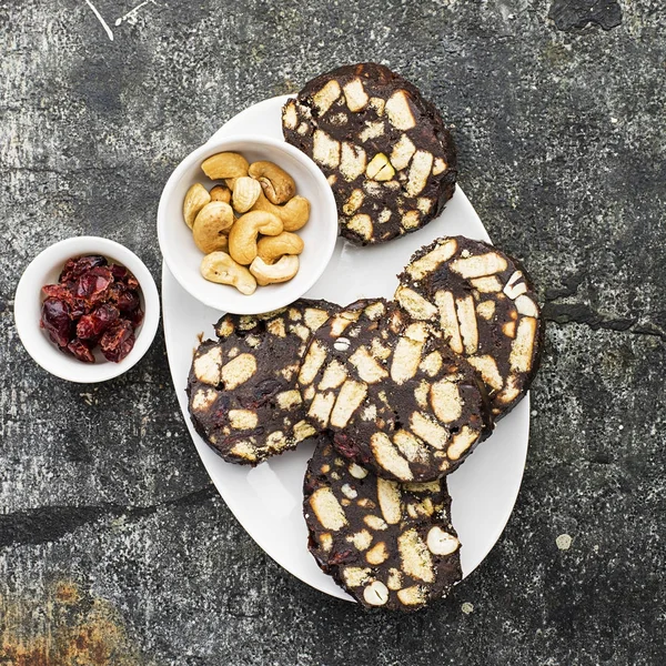 Salami de chocolate con nueces y arándanos secos servido porción sobre un fondo grunge gris con rodajas de naranja y cubiertos. Vista superior . —  Fotos de Stock