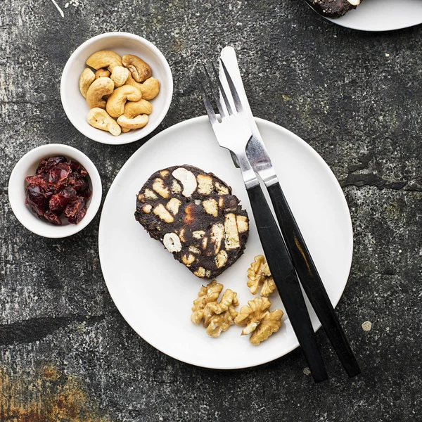 Choklad salami med nötter och torkade tranbär serveras portion på en grå grunge bakgrund med apelsinskivor och bestick. Ovanifrån. — Stockfoto