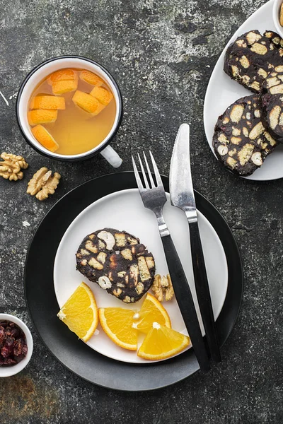 Choklad salami med nötter och torkade tranbär serveras portion på en grå grunge bakgrund med apelsinskivor och bestick. Ovanifrån. — Stockfoto