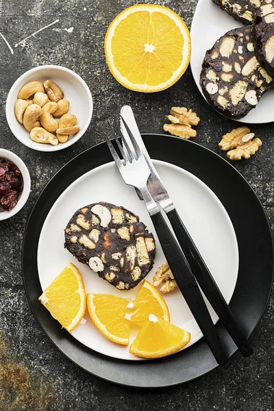 Choklad salami med nötter och torkade tranbär serveras portion på en grå grunge bakgrund med apelsinskivor och bestick. Ovanifrån. — Stockfoto