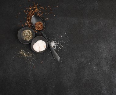 İki küçük seramik siyah el yapımı tencere ile pembe tuz ve siyah toz biber siyah yemek kaşığı ile kahverengi pirinç siyah bir arka plan ile. Üstten Görünüm. kopya alanı.