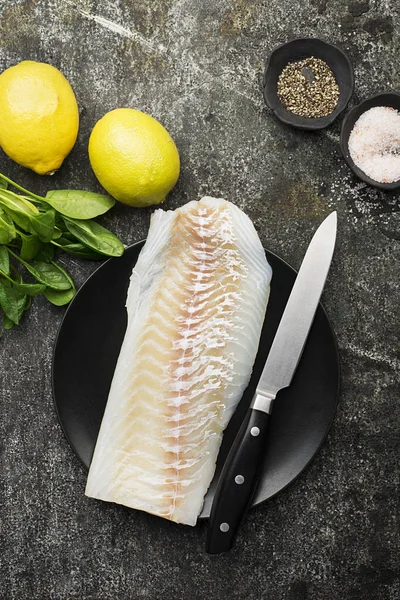 Filé de bacalhau marinho fresco para preparar um prato saudável com sal, pimenta moída, verduras sazonais frescas, fatias de limão no fundo com uma faca de cozinha para cortar. Espaço de cópia . — Fotografia de Stock