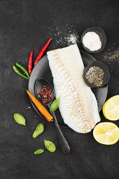 Filetes de bacalhau em uma placa de cerâmica preta para a preparação de um prato saudável com a adição de pimenta rosa, pimenta, espinafre, limão, sal em um simples fundo preto. Vista superior. Espaço de cópia . — Fotografia de Stock