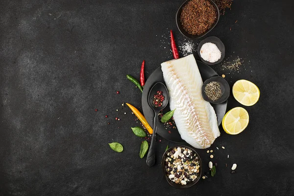 Filetes de bacalhau em uma placa de cerâmica preta para a preparação de um prato saudável com a adição de pimenta rosa, pimenta, espinafre, limão, sal em um simples fundo preto. Vista superior. Espaço de cópia . — Fotografia de Stock