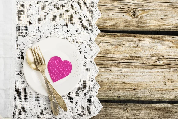 Witte keramische platen op een eenvoudige houten achtergrond met een witte vintage kanten tafelkleed met zilverwerk tabel instrumenten met een roze papieren hart. Bovenaanzicht. Kopiëren van ruimte, — Stockfoto