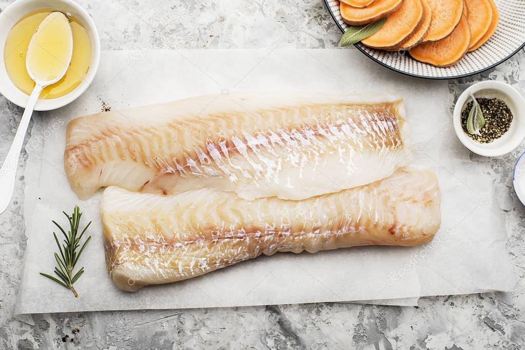 Fillet of fresh raw sea cod on a sheet of parchment on a simple light background with sea salt, ground pepper, sweet onion rosemary and lime slices before cooking. Top View.