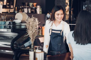 Asyalı kadın barista kahve fincanı için hizmet tutan jean önlük giymek